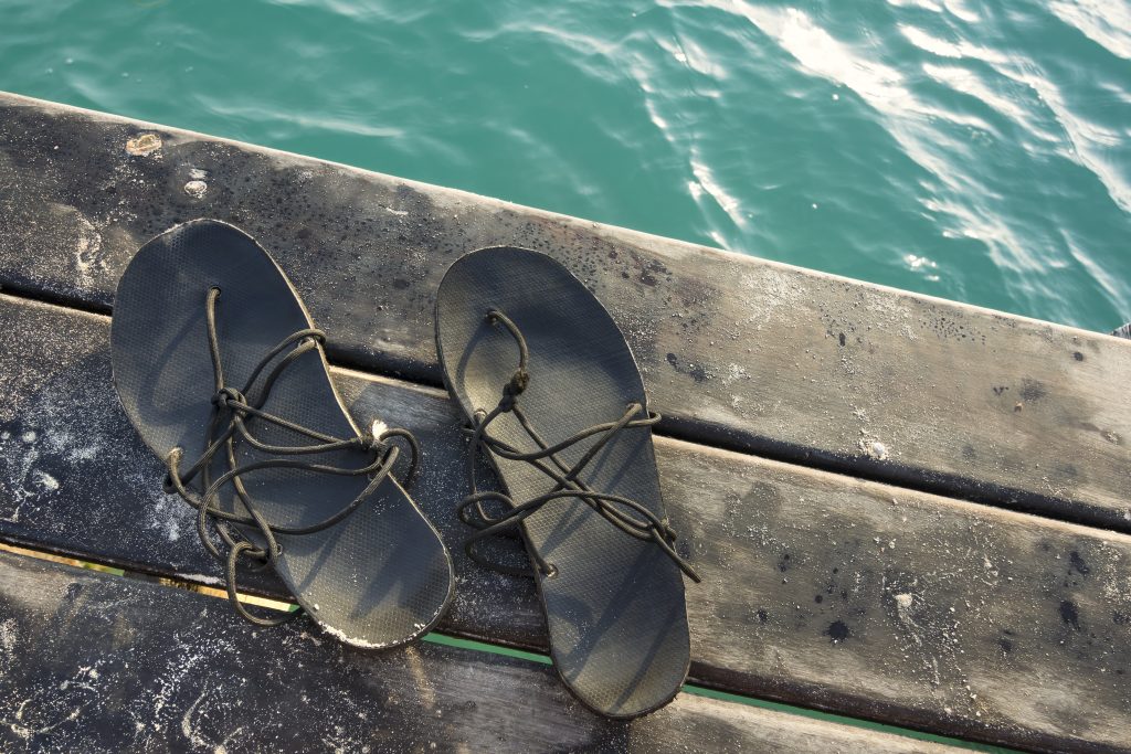 High angle view of simple black primitive style sandals on edge of wooden dock scattered with fine sand next to blue green water illuminated by morning sun